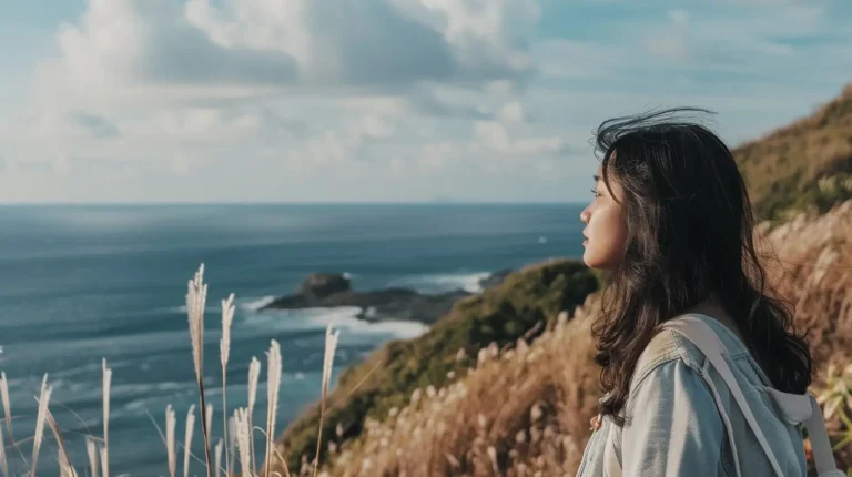 A young woman with long dark hair stands amidst tall grass on a hillside, gazing thoughtfully at the ocean. She is dressed in a light-colored jacket and carries a backpack. The sky is partly cloudy, and the blue sea stretches out to the horizon with rocky outcrops visible in the distance, perhaps considering her next adventure guided by an AI travel planner. - easytrip.ai