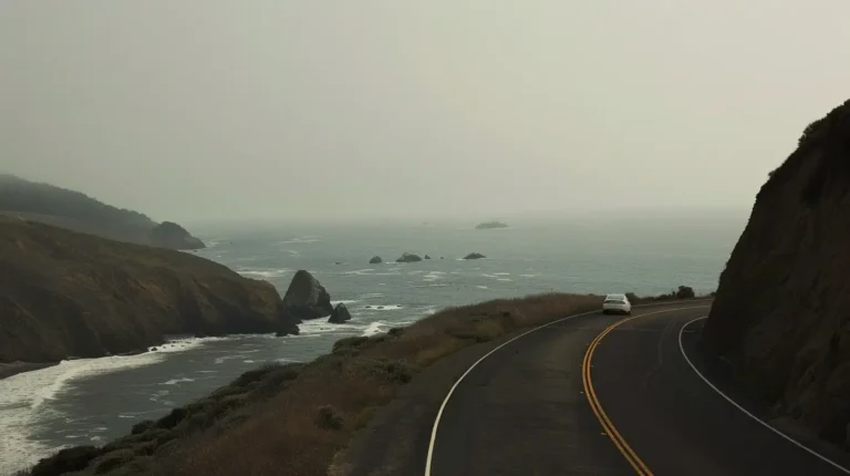 A solitary white car drives along a winding coastal road, hugging the curves of a cliff. The overcast sky creates a misty atmosphere above the rugged coastline and rocky hills. Waves crash against the shore of the grey-blue ocean in the background, stretching to the hazy horizon—an ideal scene for an AI trip planner. - easytrip.ai