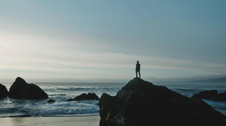 A person stands alone on a large rocky outcrop at the edge of the ocean, facing the calm waves. With an expansive sky above featuring a gradient from light to darker blue and thin, wispy clouds, the serene scene evokes solitude and contemplation—an ideal moment captured by an AI travel planner. - easytrip.ai