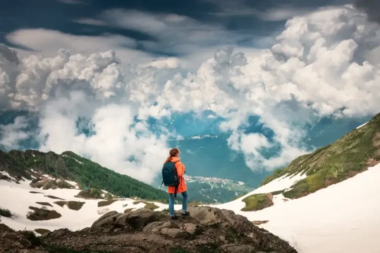 A person wearing an orange jacket and a blue backpack stands on a rocky outcrop, looking at a scenic mountain view. Snow patches dot the green mountains, and dense clouds fill the sky, creating a dramatic backdrop. With help from an AI travel planner, they'll explore more hidden gems in the valley below. - easytrip.ai