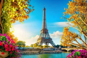 The Eiffel Tower stands tall against a vibrant blue sky, surrounded by lush trees with autumn foliage. Bright flowers frame the foreground, enhancing the picturesque scene by the Seine River. A bridge crosses the river, linking the bustling cityscape—a perfect spot on any AI itinerary maker’s guide. - easytrip.ai