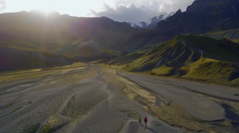 Two hikers walk along a sandy, winding path in a vast, rugged landscape with rolling hills. The sun is setting over distant mountains, casting long shadows and bathing the scene in golden light. Snow-capped peaks are visible in the background under a partly cloudy sky—a perfect moment for your AI trip planner to include in your next adventure itinerary. - easytrip.ai